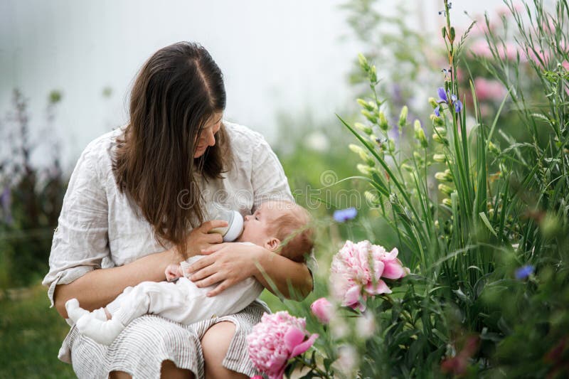 Grattis Mothers Day! Mor kramar och matar söt nyfödd med flaska i trädgården. Kärleksvård och familj på landsbygden arkivfoto