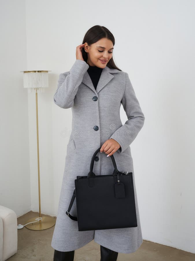 Grau Mantel mit Knöpfen getragen von einer lächelnden Frau, die schwarze Handtasche im Zimmer mit Bodenlampe und Sofa hält. Sie ha stockbild