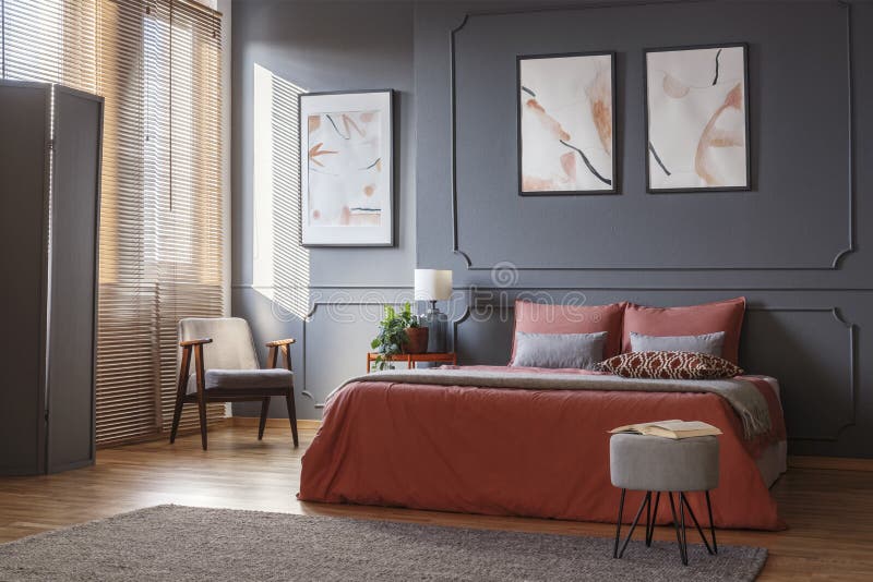 Gray, retro armchair standing in the corner of an elegant bedroom interior with watercolor posters on dark gray wall with molding and orange sheets on the bed. Real photo. Gray, retro armchair standing in the corner of an elegant bedroom interior with watercolor posters on dark gray wall with molding and orange sheets on the bed. Real photo