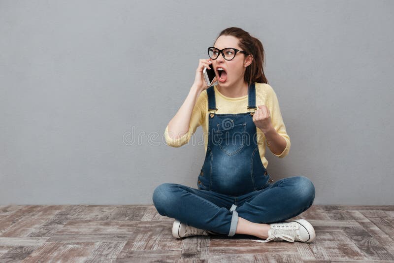 Image of pregnant angry screaming woman wearing glasses sitting isolated over grey wall. Talking by mobile phone. Image of pregnant angry screaming woman wearing glasses sitting isolated over grey wall. Talking by mobile phone.