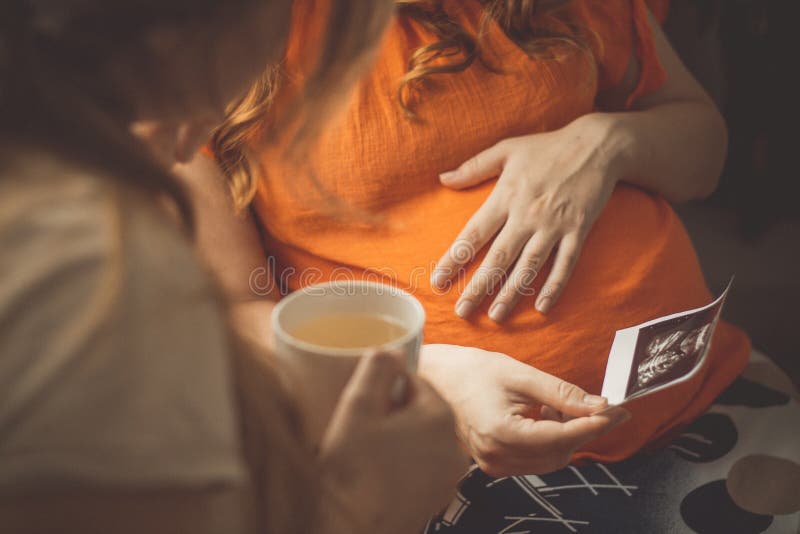 Gravid ung kvinna som sitter i köket med sistertal om ultraljudsbilder moderskapskoncept graviditet och vänskap royaltyfri bild