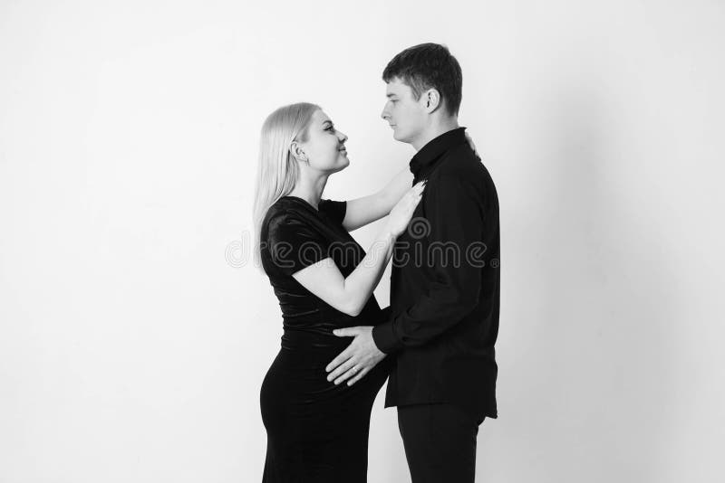 Gravida kvinnor och hennes make kramar och håller fast vid magen i svarta kläder på vit bakgrund Svartvit fotografering för bildbyråer