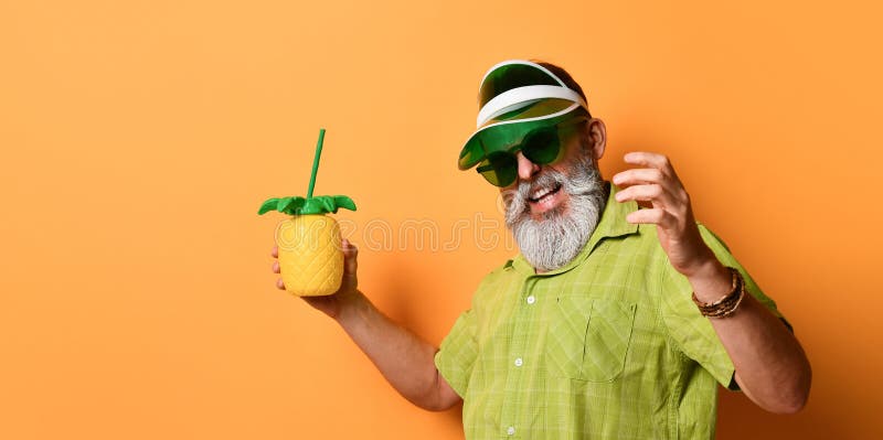 Grandpa in green sun visor, shirt, sunglasses. Holding cocktail bottle in form of pineapple with tube, posing on orange background. Gray-bearded grandpa in green stock photos Grandpa in green sun visor, shirt, sunglasses. Holding cocktail bottle in form of pineapple with tube, posing on orange background. Gray-bearded grandpa in green stock photos