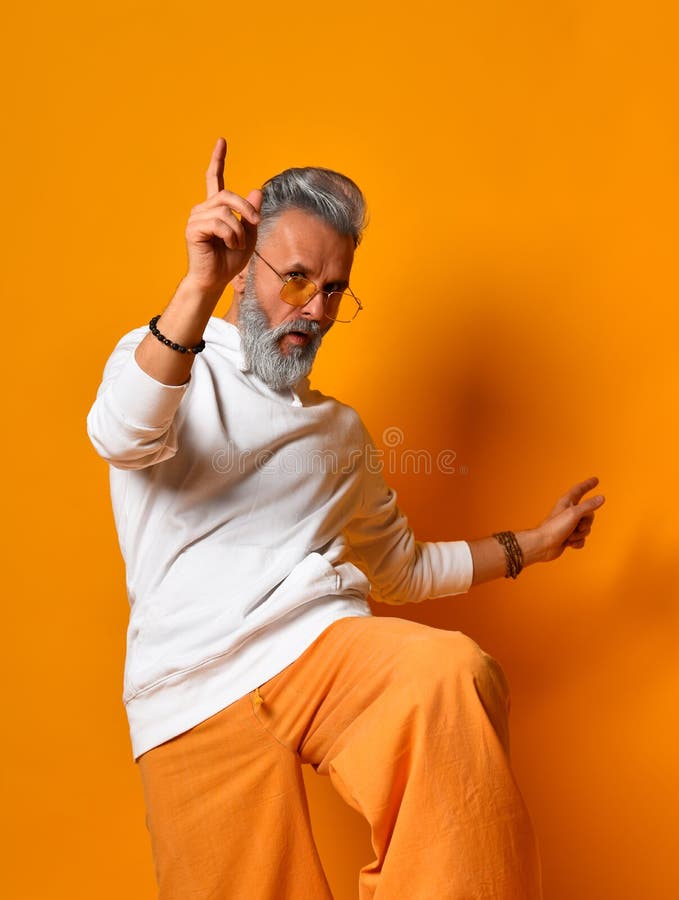 Gray-haired granddad in white hoodie, pants and sunglasses, bracelets. Raised his leg up, posing on orange background. Close up. Gray-haired, bearded granddad in stock images Gray-haired granddad in white hoodie, pants and sunglasses, bracelets. Raised his leg up, posing on orange background. Close up. Gray-haired, bearded granddad in stock images
