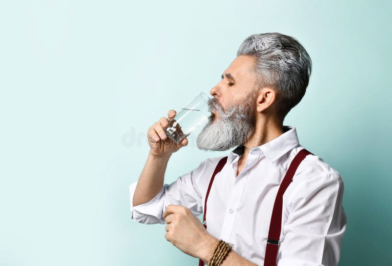 Gray-haired elderly male in white shirt, brown suspenders. He drinking pure water from glass, posing sideways on blue background. Gray-haired, bearded elderly stock photos Gray-haired elderly male in white shirt, brown suspenders. He drinking pure water from glass, posing sideways on blue background. Gray-haired, bearded elderly stock photos