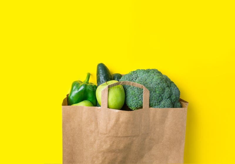 Green raw organic vegetables fruits broccoli cucumbers bell peppers apples in brown paper Kraft grocery bag on yellow background