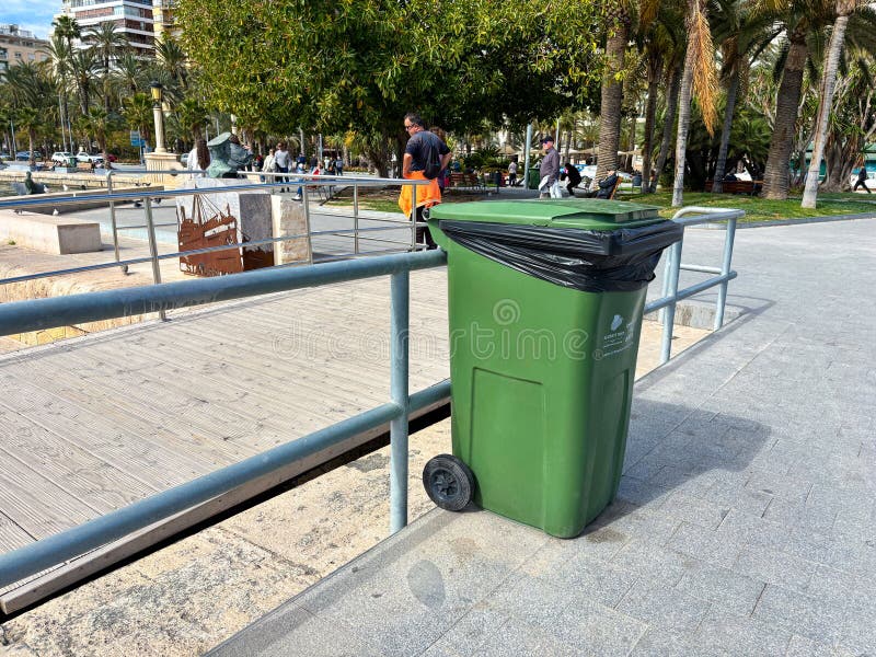Green trash bin positioned near a park walkway with people in the background, promoting cleanliness and environmental awareness royalty free stock photography
