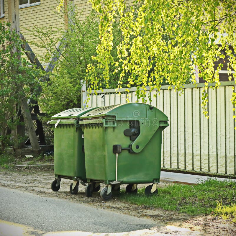 Green trash containers in sunlit street near fence with birch tree branches and greenery royalty free stock photography