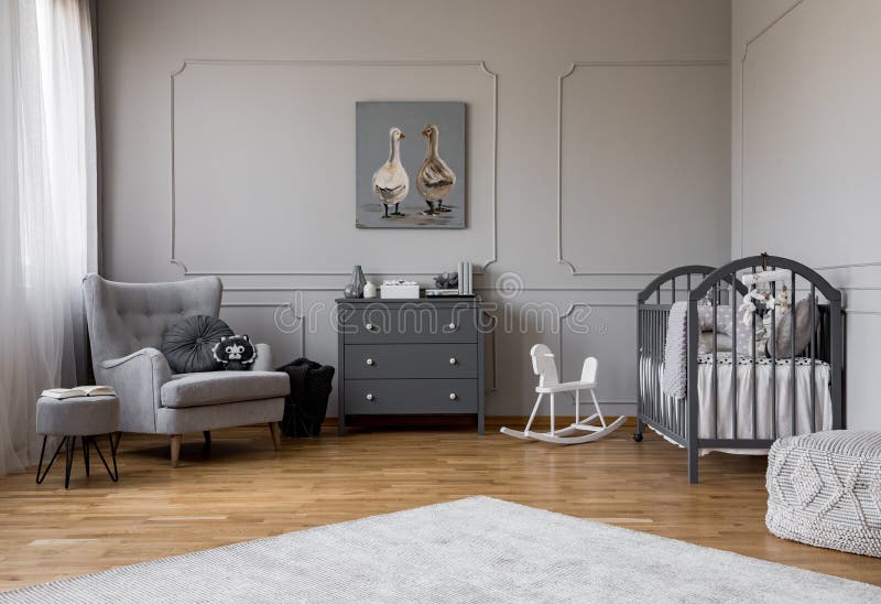 Grey chest of drawers in the middle of elegant grey baby room with comfortable armchair and wooden cradle