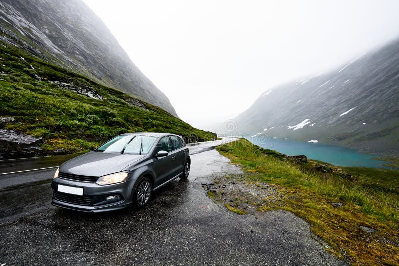 Grey modern car is parking next to a rural road in a valley surrounded by a fjord and snow covered mountains on a rainy day in Nor