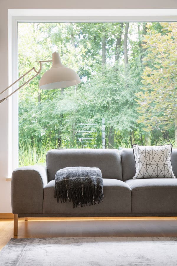 Grey settee with patterned cushion and blanket standing in real photo of sitting room interior with carpet, oversize lamp and big