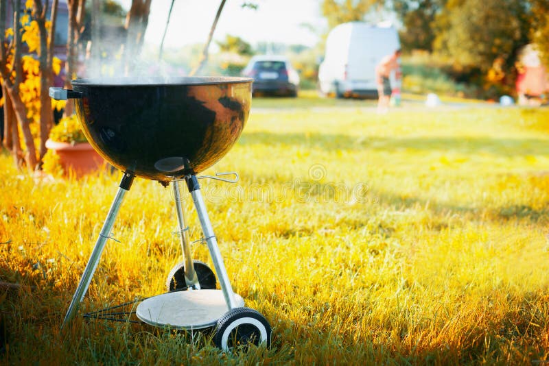 Grill with smoke at late summer or autumn nature background in a park or garden with silhouettes of cars and people