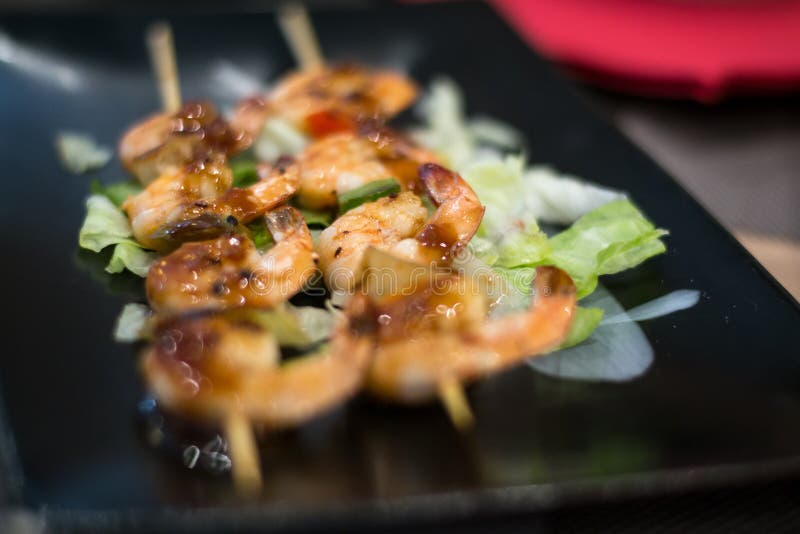 A black plate of grilled scrimp on a wooden spike, covered with some barbecue sauce and with a little salad as dressing. A black plate of grilled scrimp on a wooden spike, covered with some barbecue sauce and with a little salad as dressing.