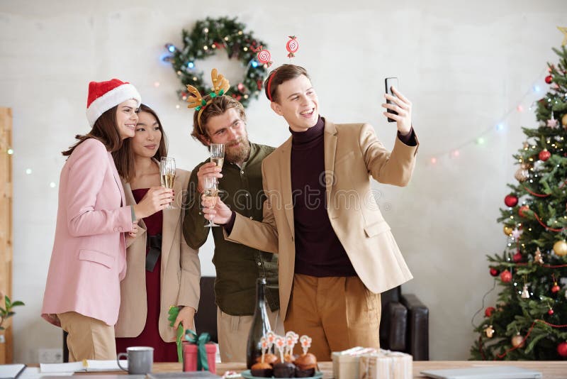 Group of young contemporary business partners with flutes of champagne looking at camera in smartphone held by elegant man during toast. Group of young contemporary business partners with flutes of champagne looking at camera in smartphone held by elegant man during toast