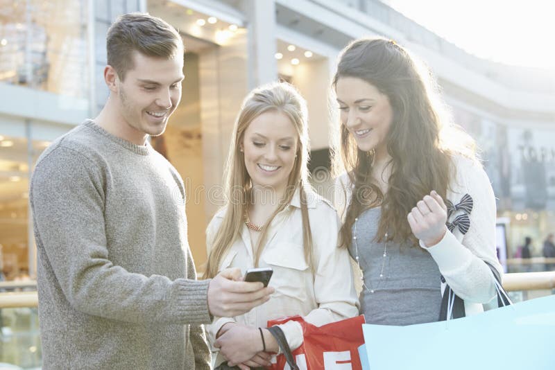 Groep Vrienden die in Wandelgalerij winkelen die Mobiele Telefoon bekijken stock fotografie Groep Vrienden die in Wandelgalerij winkelen die Mobiele Telefoon bekijken stock fotografie