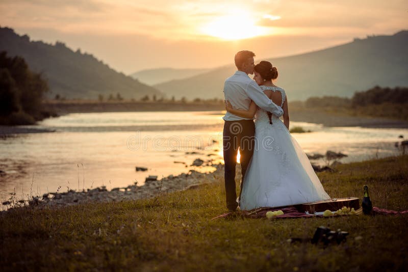 Groom is softly kissing his gorgeous bride in the forehead during the sunset. Wedding picnic on the river bank