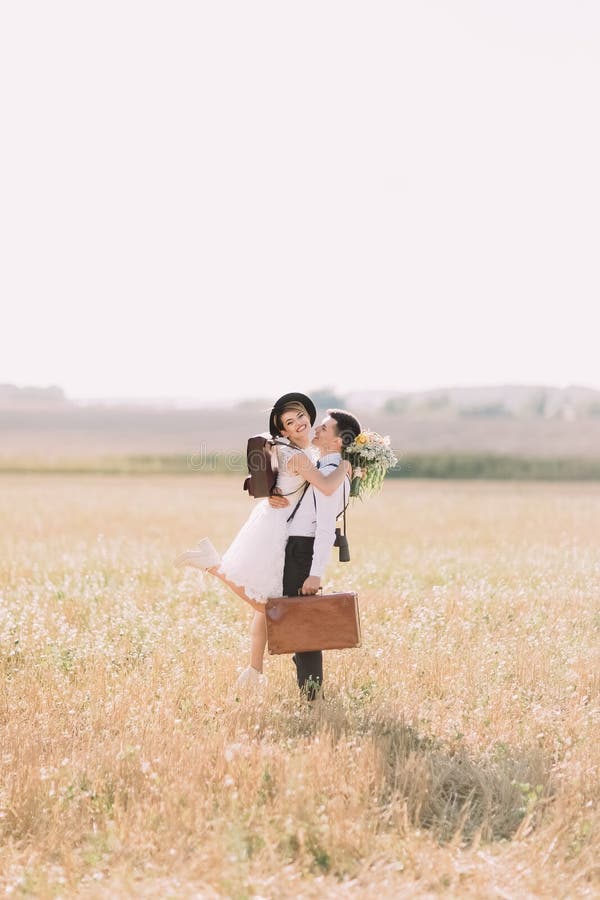 The groom with the vintage suitcase is holding the smiling bride in the arms at the background of the sunny field.