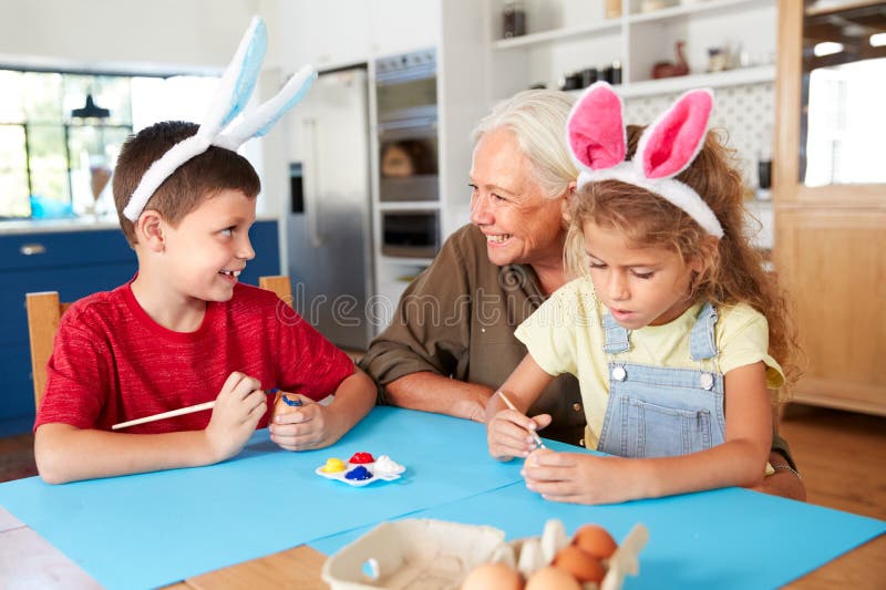 Grootmoeder met Kleinkinderen die Konijnoren dragen die Paaseieren thuis samen verfraaien royalty-vrije stock foto's Grootmoeder met Kleinkinderen die Konijnoren dragen die Paaseieren thuis samen verfraaien royalty-vrije stock foto's