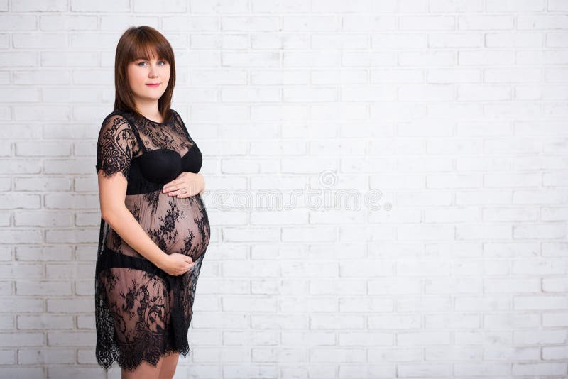 Grossesse - femme dans la robe noire de dentelle posant au-dessus du wa blanc de brique images stock Grossesse - femme dans la robe noire de dentelle posant au-dessus du wa blanc de brique images stock