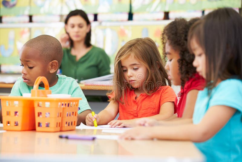 Group Of Elementary Age Children In Art Class With Teacher Sitting At Table Drawing