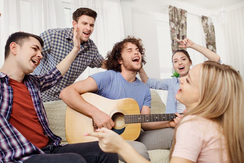 A group of friends with a guitar sing fun songs at a party indoor.