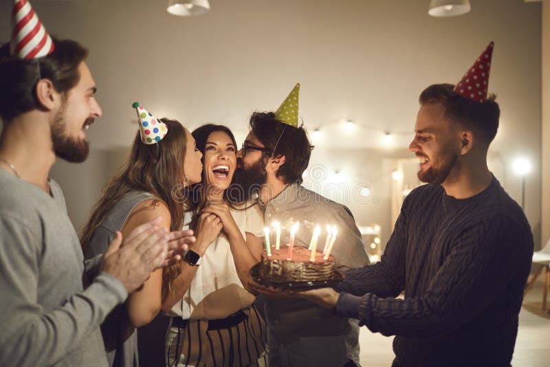 Group of happy, cheerful young people congratulating their friend on her birthday, kissing her on cheeks and giving her a chocolate cake with burning candles during a party at home. Group of happy, cheerful young people congratulating their friend on her birthday, kissing her on cheeks and giving her a chocolate cake with burning candles during a party at home