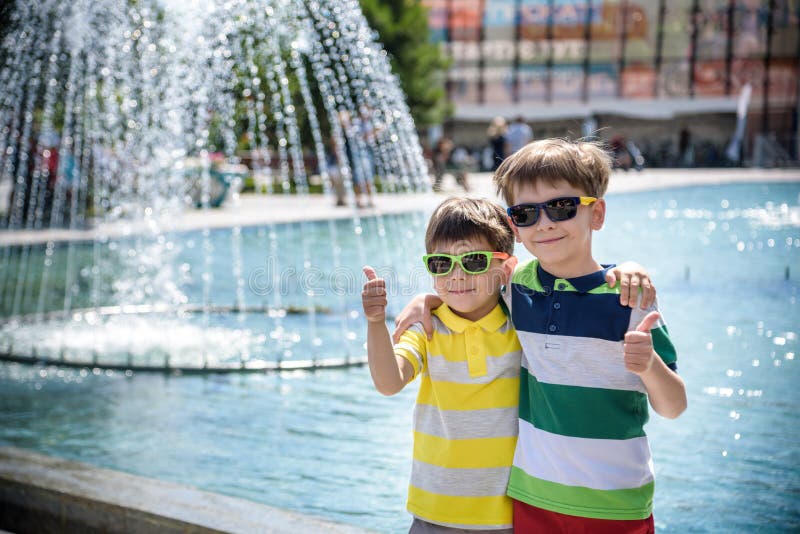 Group of happy children playing outdoors near pool or fountain. Kids embrace show thumb up in park during summer vacation. Dressed in colorful t-shirts and shorts with sunglasses. Summer holiday concept. Group of happy children playing outdoors near pool or fountain. Kids embrace show thumb up in park during summer vacation. Dressed in colorful t-shirts and shorts with sunglasses. Summer holiday concept