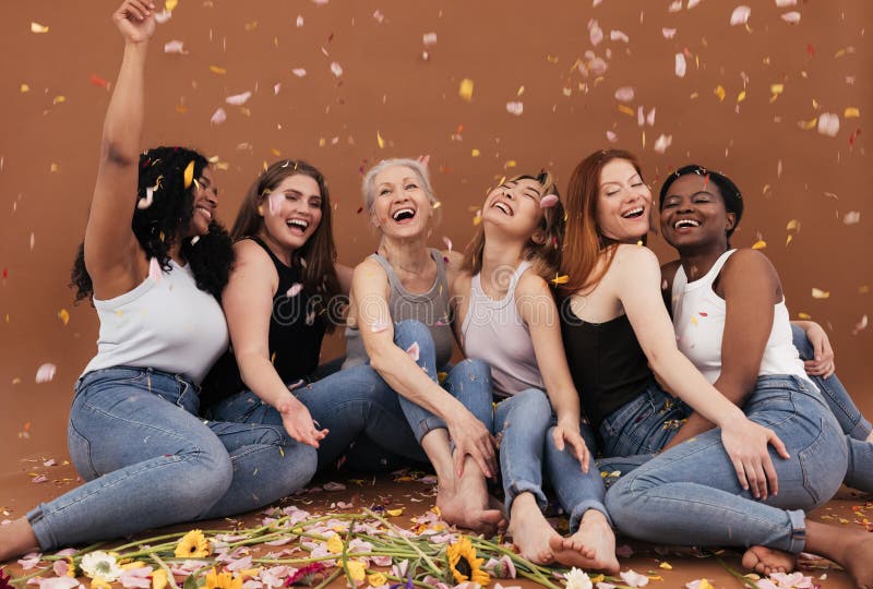 Group of happy women sitting on the brown background while petals from flowers falling. Laughing females of different races and