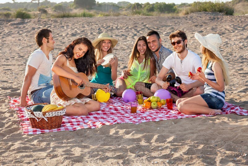 Group of young people having an enjoyable picnic on the beach with healthy food, three of them playing music, young female is playing guitar. Group of young people having an enjoyable picnic on the beach with healthy food, three of them playing music, young female is playing guitar.