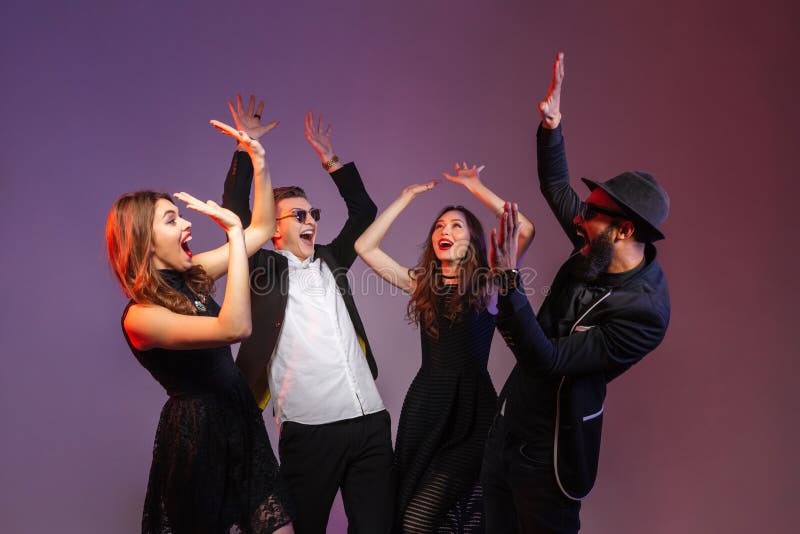 Group of happy young people standing together with raised hands over purple background