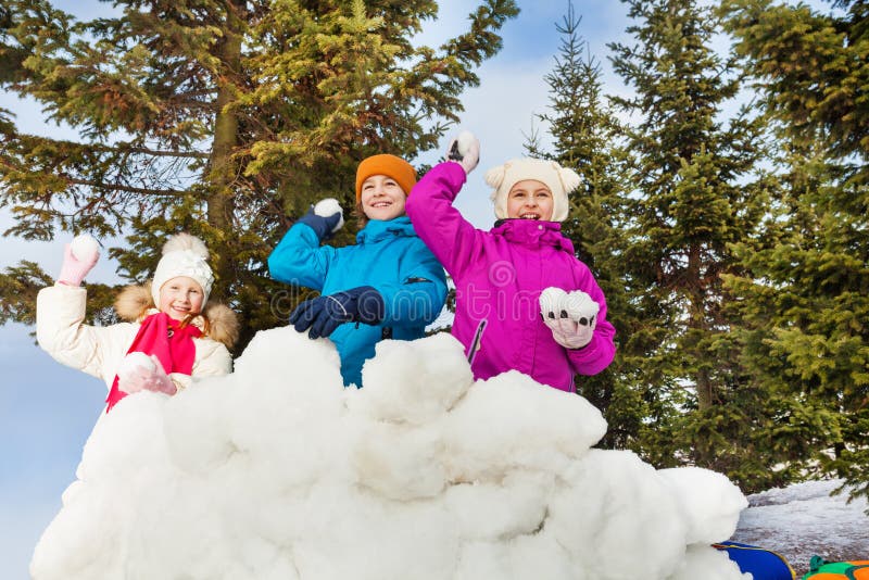 Group of kids play snowballs game together. Standing behind the snow wall fortress with fir forest on the background during winter day stock images