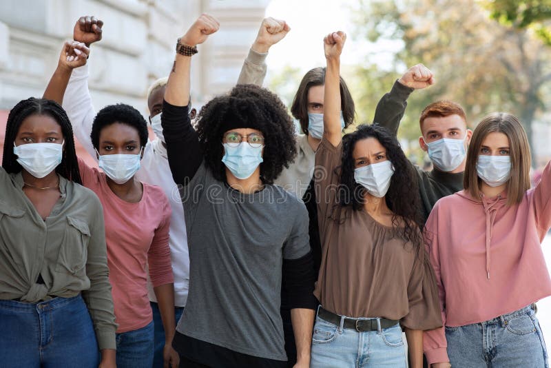 Multiracial group of young people wearing face mask protesting and giving slogans on the street. Group of demonstrators protesting in the city during quarantine, raising hands up and yelling. Multiracial group of young people wearing face mask protesting and giving slogans on the street. Group of demonstrators protesting in the city during quarantine, raising hands up and yelling