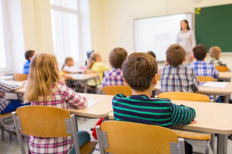 Education, elementary school, learning and people concept - group of school kids sitting and listening to teacher in classroom from back. Education, elementary school, learning and people concept - group of school kids sitting and listening to teacher in classroom from back