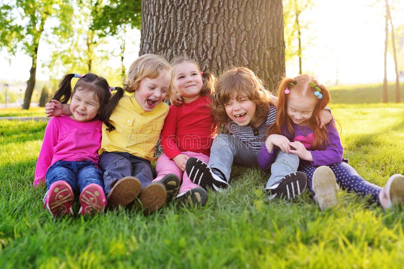 A group of small children in colorful clothes embracing sitting on the grass under a tree in a park laughing and smiling royalty free stock image A group of small children in colorful clothes embracing sitting on the grass under a tree in a park laughing and smiling royalty free stock image