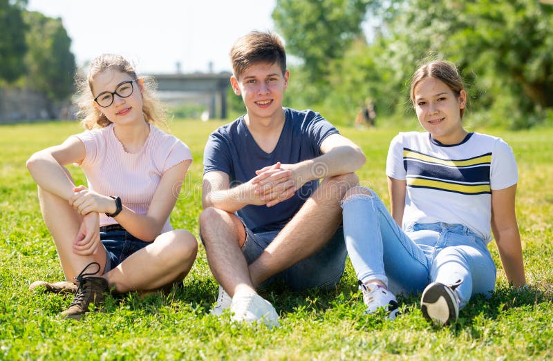 Smiling teenage boy and girls sitting on green grass. Group of smiling teenage boy and two girls having fun together on summer day, sitting on green grass