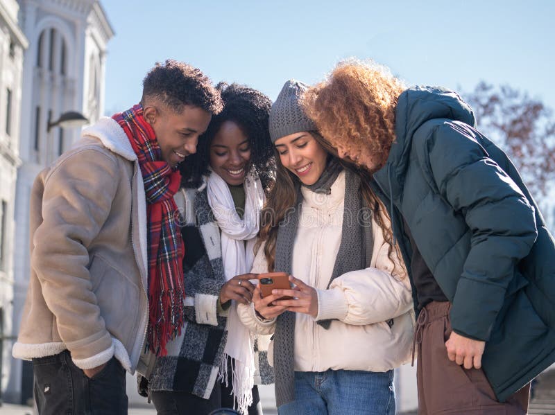Low angle view of four cheerful multiethnic students using a smartphone together outdoors in winter royalty free stock photo