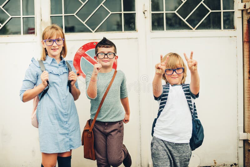 Group of three funny kids wearing backpacks walking back to school. Girl and boys wearing eyeglasses posing outdoors