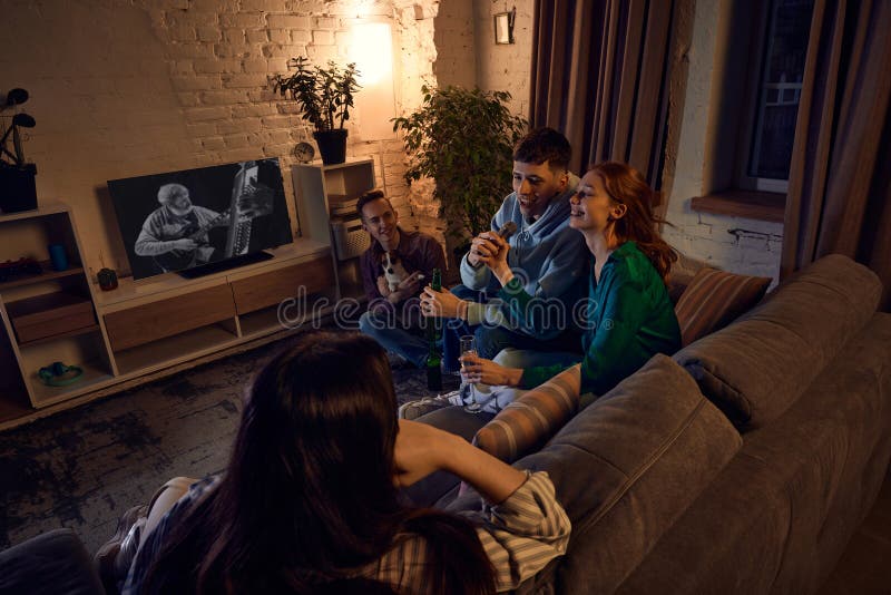 Group of young, cheerful people, friends sitting on couch at home in the evening and singing karaoke, talking, drinking