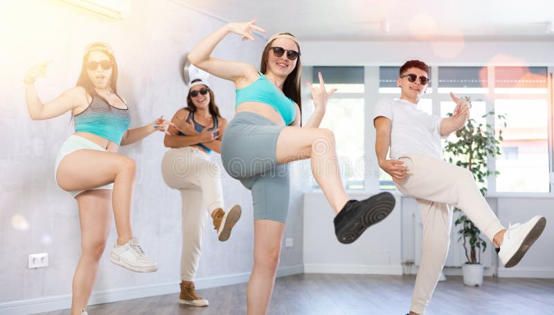 Female teenager performs movements during warm-up, limbering-up part of workout together with peers. Guy and group of young girls in dark glasses and ball cap dance jazz-funk in fitness club. Female teenager performs movements during warm-up, limbering-up part of workout together with peers. Guy and group of young girls in dark glasses and ball cap dance jazz-funk in fitness club.