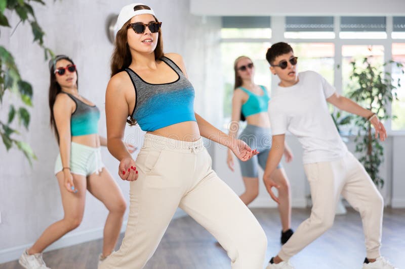 Female teenager performs movements during warm-up, limbering-up part of workout together with peers. Guy and group of young girls in dark glasses and ball cap dance shuffle in fitness club. Female teenager performs movements during warm-up, limbering-up part of workout together with peers. Guy and group of young girls in dark glasses and ball cap dance shuffle in fitness club.