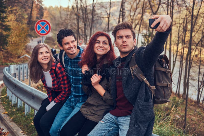 Group of young hikers with backpacks sitting on guardrail near road and doing selfie. stock images Group of young hikers with backpacks sitting on guardrail near road and doing selfie. stock images