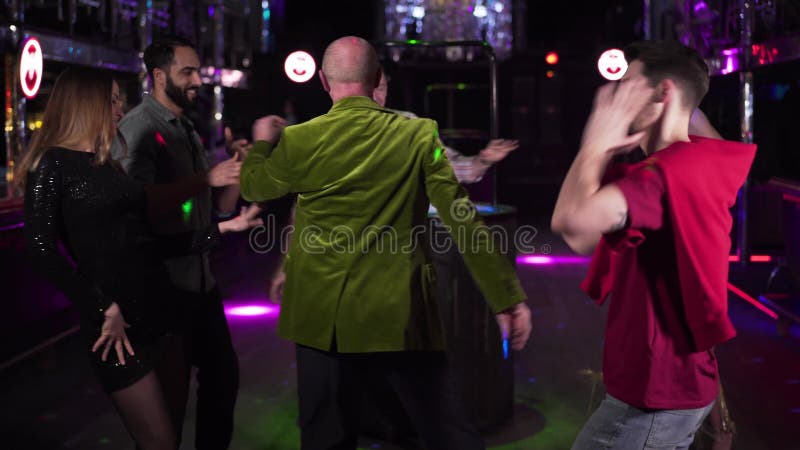 Group of young multi-ethnic people dancing around cheerful elderly man in night club. Positive men and women resting in