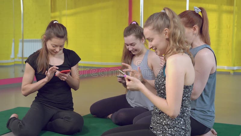 Group of young women in sportswear actively using phones. Four girl sitting on the mat totally focused on their smartphones