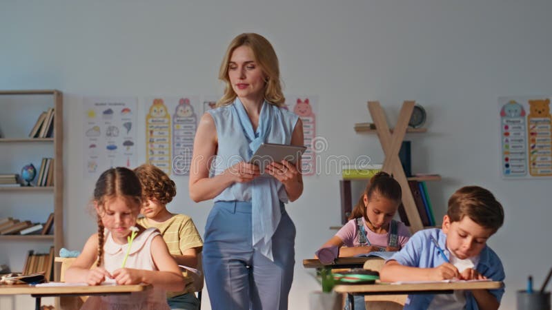 Groupe d'enfants faisant des devoirs, des tables de classe. Une jeune enseignante regarde clips vidéos