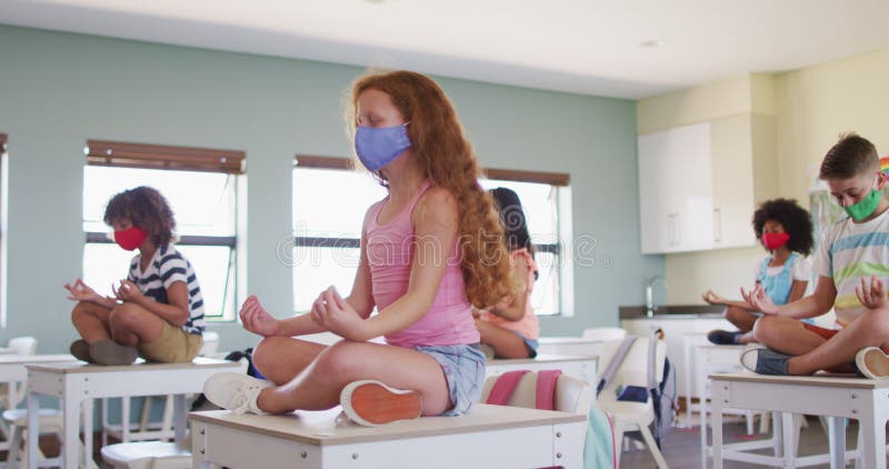Groupe d'enfants portant des masques de protection pratiquant le yoga tout en reposant sur des bureaux à l'école banque de vidéos