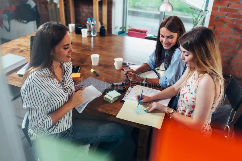 Groupe de femmes créatives de sourire discutant un projet se reposant autour de la table faisant des notes dans le bureau photographie stock libre de droits Groupe de femmes créatives de sourire discutant un projet se reposant autour de la table faisant des notes dans le bureau photographie stock libre de droits