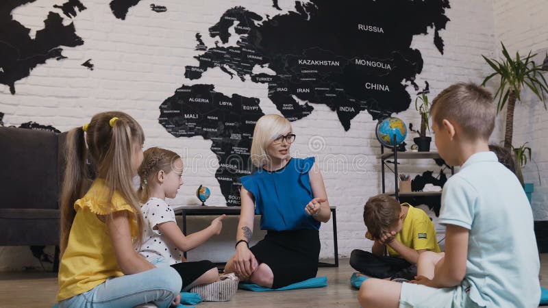 Groupe de petits élèves de cinq enfants s'asseyant sur le plancher et écoutant le professeur Enfants et éducation, jeune blonde