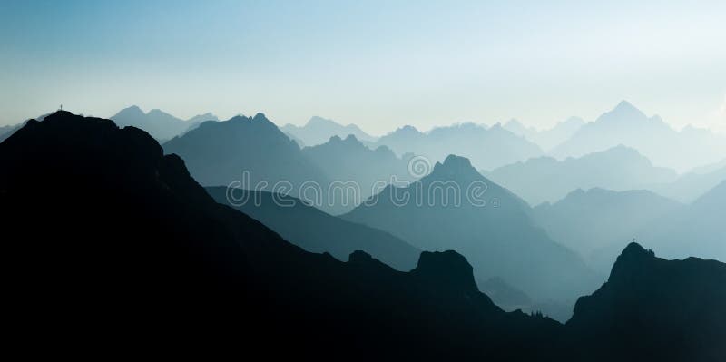 Blue and cyan mountain ranges silhouette with bright back light. Tirol, Austria. Summit crosses are visible from afar on some mountain peaks. Blue and cyan mountain ranges silhouette with bright back light. Tirol, Austria. Summit crosses are visible from afar on some mountain peaks.