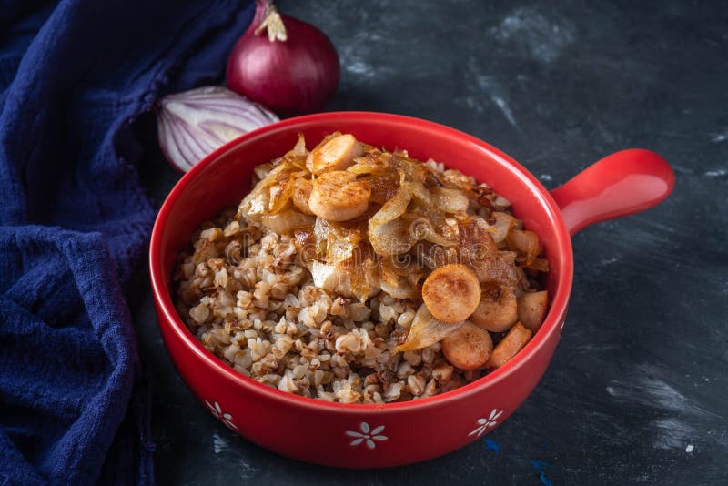 Gruau de sarrasin avec des saucisses et des oignons frits dans une casserole rouge sur un fond foncé, l'espace de copie image libre de droits Gruau de sarrasin avec des saucisses et des oignons frits dans une casserole rouge sur un fond foncé, l'espace de copie image libre de droits