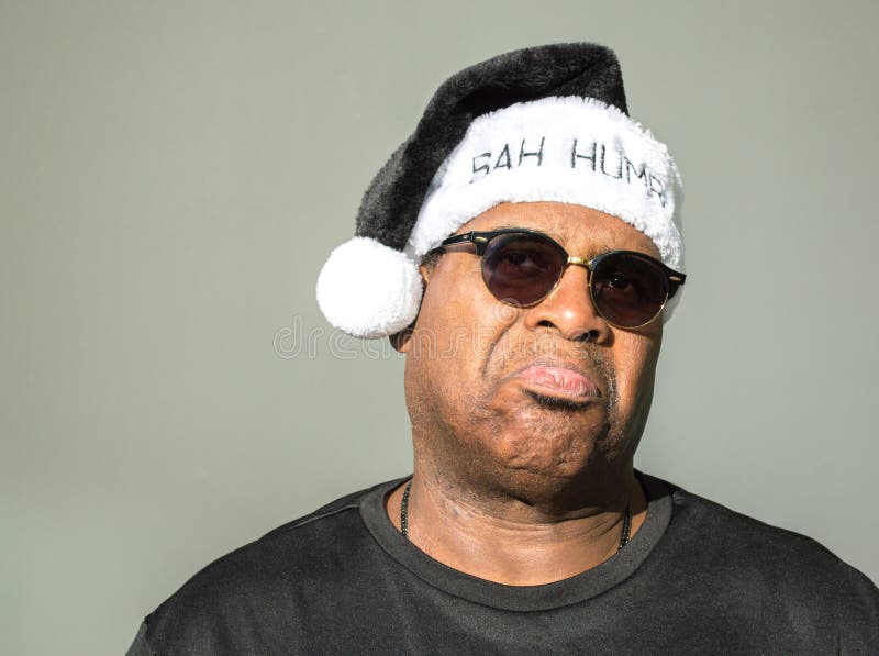 Cranky middle aged rotund African American man wearing a black and white Santa hat saying Bah Humbug against a solid background. Cranky middle aged rotund African American man wearing a black and white Santa hat saying Bah Humbug against a solid background